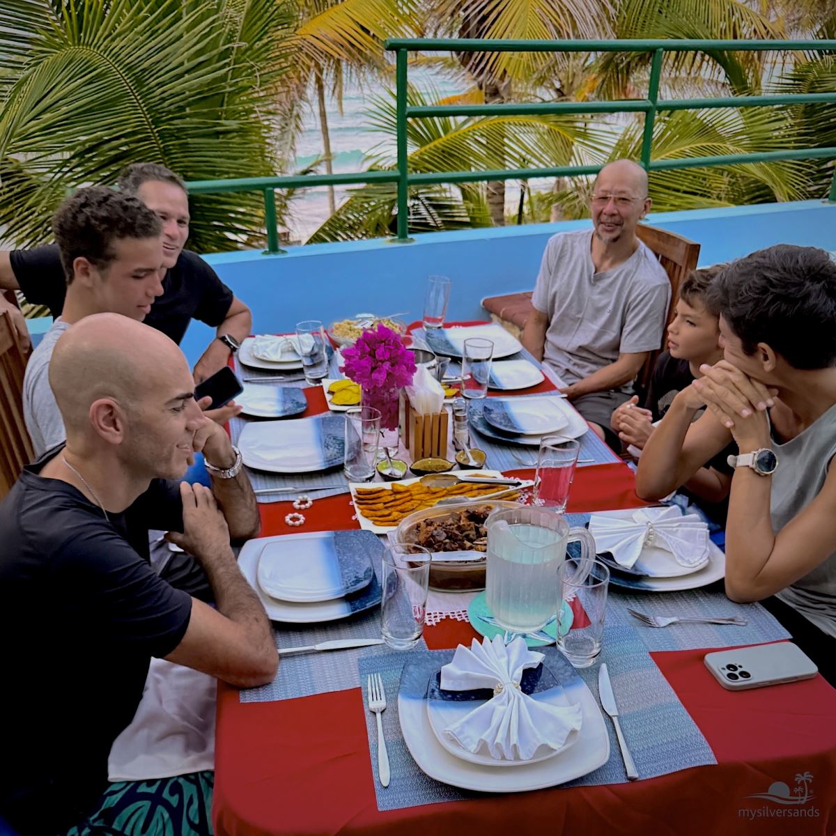 Friends and family gathered for dinner on a veranda at Silver Sands with palm trees and the sea in the background.